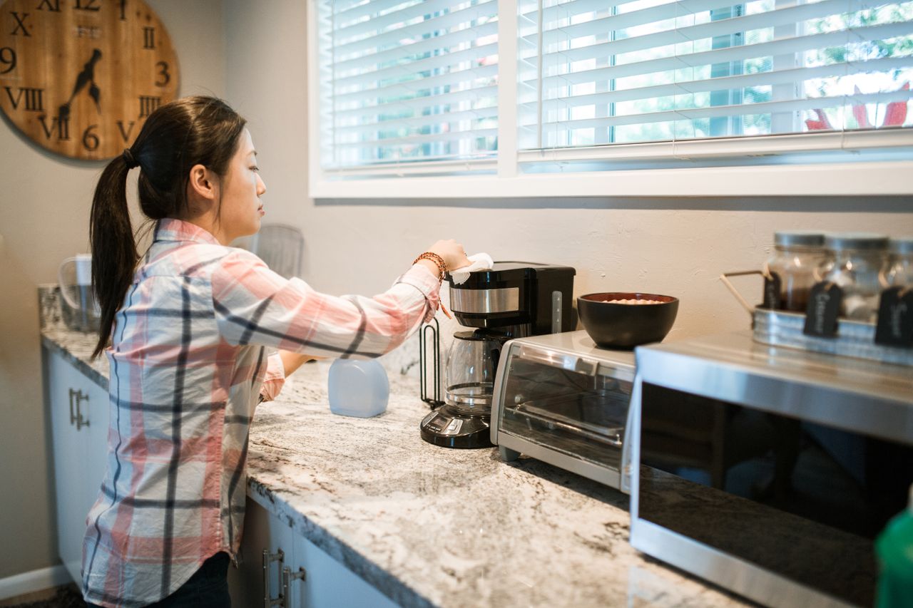 who-we-are Woman cleaning a coffee maker in a bright, modern kitchen with appliances.